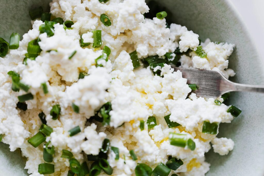 Top view of gray bowl with meal cottage cheese with pieces of green onion and fork placed on kitchen meal for breakfast